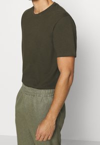 Tumman oliivinvihreä puuvillapaidan t-paita, jossa on pyöreä kaula-aukko, lyhyet hihat ja rento malli. Yhdistetty vaaleanharmaisiin, teksturoituihin housuihin.