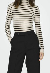 Femme portant un pull à col roulé côtelé à rayures crème et noires, avec un pantalon noir taille haute, une main dans la poche et l'autre détendue.
