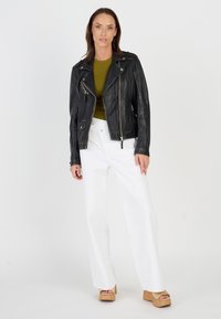 Schwarze Leder-Motorradjacke mit silbernen Reißverschlüssen, olivgrünes Oberteil, weiße weite Hose und braune Plateauschuhe mit Korkverzierungen.
