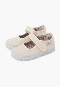 Scarpe Mary Jane in tela color sabbia con una fascetta in velcro, suole in gomma bianca e una fodera interna a quadretti marroni e bianchi.