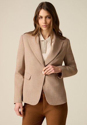 Kobieta z długimi brązowymi włosami, ubrana w beżową marynarkę na kremowej koszuli z perłowymi zdobieniami, zapinająca guzik marynarki, stoi na jednolitym tle.