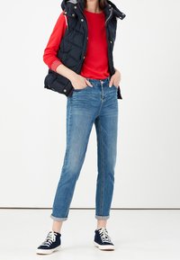 Person som bär en röd långärmad skjorta, en svart vadderad väst, blå uppvikta jeans och navy höga sneakers står med händerna i fickorna.