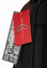 Mochila negra con una superficie texturizada, que presenta una gran etiqueta roja con la inscripción "SPRAYGROUND" y un diseño impreso de billete de dólar.