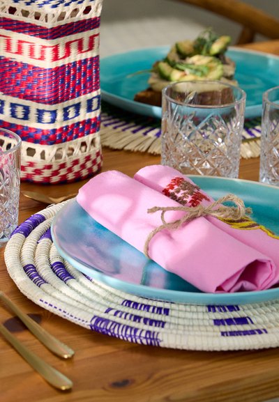 Pink cloth napkins tied with twine on a blue plate, layered on a round woven placemat. Crystal glasses and a patterned basket are in the background.