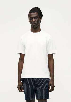 Homme noir avec des dreadlocks porte un t-shirt blanc uni à manches courtes et un short bleu marine avec des rayures blanches verticales, debout devant un fond uni.