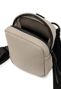 Piccola borsa a tracolla beige con superficie liscia, chiusura con zip, tracolla nera e tasca interna. Presenta cuciture sottili e logo.