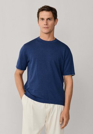 Hombre con camiseta azul marino de manga corta y cuello redondo y pantalones beige claro, de pie con una mano en el bolsillo contra un fondo liso.