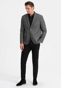 Blazer gris texturizado con solapa de pico, dos bolsillos frontales, cierre de un botón, combinado con camisa negra y pantalones negros ajustados.