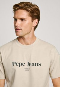 Bēša kokvilnas T-krekls ar "Pepe Jeans" logo tumši zaļā krāsā, apakšā zem galvenā logo ir papildu teksts par atrašanās vietu mazākā fontā.