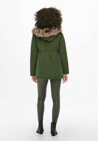 Parka verte avec capuche bordée de fourrure, taille cintrée et manches longues. Portée avec des leggings verts brillants et des bottines. Fond blanc.