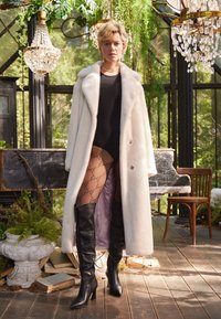 Weißer Kunstpelzmantel mit Schalkragen, schwarzes Body, Netzstrumpfhosen mit Logo-Muster und knielange schwarze Stiefel mit spitzer Zehe.