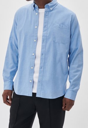 Mand iført en lyseblå skjorte med knapper over en hvid t-shirt og sorte bukser, vist fra nakke til hofter.