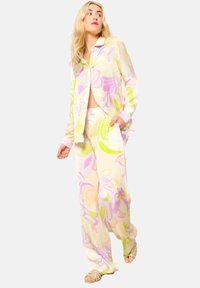 Lange mouwen blouse met knopen en losse broek van doorschijnende stof, met een pastelbloemenpatroon in paarse en groene tinten op een lichtgele achtergrond.
