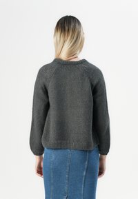 Žena s rovnými blond vlasy, která má na sobě tmavě šedý pletený svetr a modrou džínovou sukni, obrácená zády k jednobarevnému bílému pozadí.