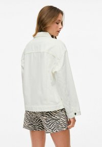 Chaqueta de mezclilla blanca con un corte relajado, que presenta un cuello y detalles de botones, combinada con pantalones cortos a rayas beige y negros.