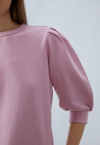 Žena nosící pastelově růžový dlouhý rukáv s nafouklými rukávy a žebrovanými manžetami, stojící před jednoduchým světlým pozadím.