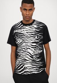 Fekete pamut póló, fehér zebra csíkos mintával, a következő szöveggel: "Versace Jeans Couture", szabálytalan betűtípussal.