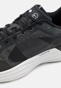 Svart sportskor med ovan del av mesh och syntetmaterial, strukturerade mönster, vadderad vit sula och framträdande Nike-logga på sidan.