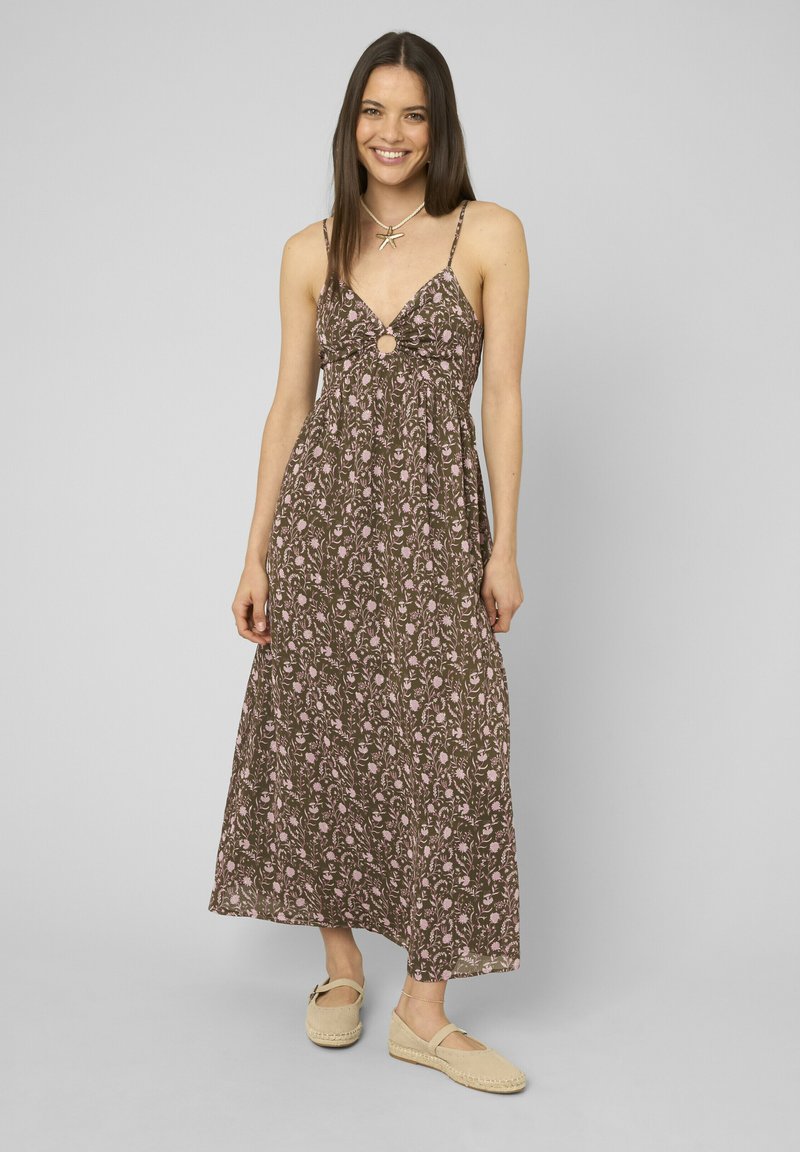 Femme souriante portant une robe maxi marron sans manches avec un motif floral rose et des espadrilles beiges, debout devant un fond uni.