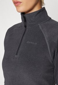 Mujer vistiendo un pulóver de forro polar acanalado gris oscuro con media cremallera y cuello alto, que presenta un sutil logo bordado en el pecho.