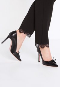 Černé kožené lodičky na vysokém podpatku s špičatou špičkou a mašlí, s elegantním designem a stiletto podpatkem, spárované s černými kalhotami zdobenými krajkou.