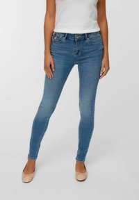 Sieviete gaiši zilos šaurajos džinsos un bēšos balerīnu apavos, stāvot pret parastu gaiši pelēku fona.