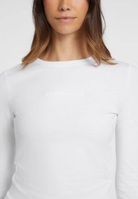 Camicia bianca a maniche lunghe con scollatura a girocollo e testo del logo in rilievo. Il tessuto sembra elastico e liscio al tatto.