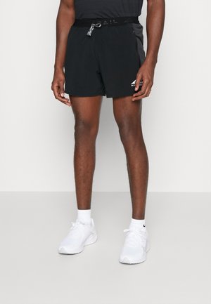 Homme portant un short de sport noir avec poches latérales et des baskets blanches, debout devant un fond blanc uni.