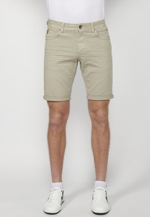 Homme portant un short beige avec ourlet roulé, associé à des chaussettes blanches et des baskets blanches, debout devant un fond uni.