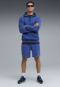 Felpa con cappuccio in denim blu con tasca frontale e cordino, abbinata a pantaloni corti blu coordinati. Completata con sneakers navy e occhiali da sole blu.