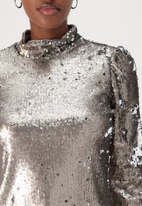 Metallisch silberner Paillettenoberteil mit hohem Kragen und Puffärmeln, der über eine glatte Textur und eine reflektierende Oberfläche verfügt.