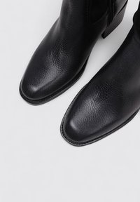 Botas de tobillo de cuero negro con superficie texturizada, puntera afilada y tacón cuadrado. Costuras visibles a lo largo de la suela para un detalle adicional.