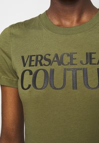Olivově zelené tričko s krátkým rukávem vyrobené z bavlny, s velkým černým potiskem loga "VERSACE JEANS COUTURE" přes hrudník.