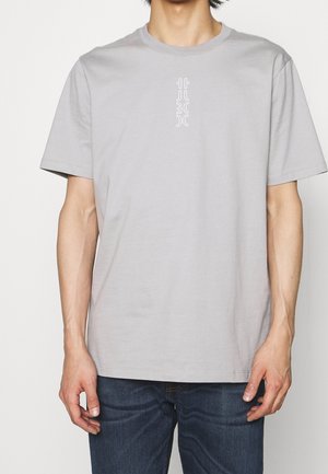 Mies, jolla on vaaleanharmaa lyhythihainen t-paita, jossa on pieni valkoinen pystysuora grafiikka rinnassa, sekä tummansiniset farkut, seisoo yksiväristä taustaa vasten.