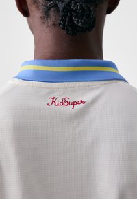 La chemise à col présente un tissu en maille crème clair, avec un col bleu accentué d'une rayure jaune. "KidSuper" est brodé en rouge.