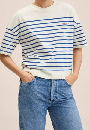 Persoon draagt een wit T-shirt met blauwe horizontale strepen en rood opschrift, licht ingestopt in een high-waisted blauwe spijkerbroek met handen in de zakken.