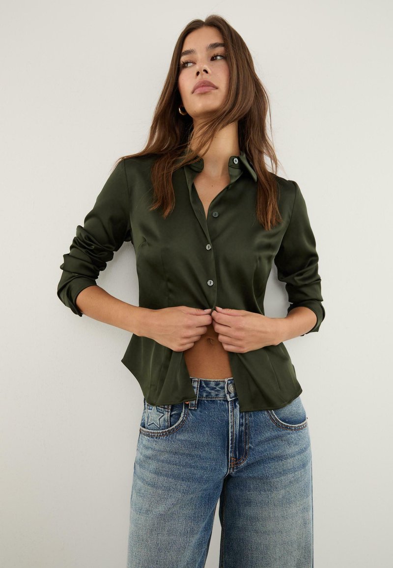 Camicia button-up in satin verde scuro con maniche lunghe, caratterizzata da un orlo curvo e una texture liscia, abbinata a jeans a gamba larga di colore azzurro chiaro.