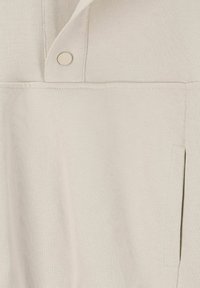 Tissu tricoté beige clair avec un col, présentant une fermeture à boutons et une poche latérale. Texture lisse, design simple avec des lignes épurées.