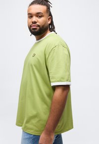 Άνδρας με πλεξούδες στα μαλλιά και γενειάδα, φορώντας ανοιχτό πράσινο μπλουζάκι με λευκή επένδυση και μπλε τζιν, στέκεται μπροστά σε απλό λευκό φόντο.