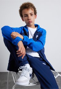 Nike trainingspak in blauw en marineblauw, met een zip-up jack en bijpassende broek. Gekleed met witte sneakers met blauwe accenten. Gladde stofstructuur.