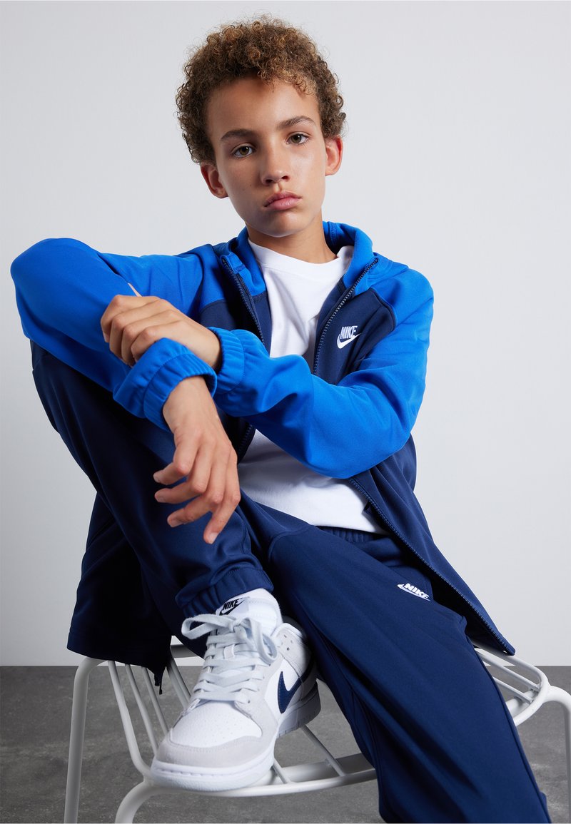 Nike trainingspak in blauw en marineblauw, met een zip-up jack en bijpassende broek. Gekleed met witte sneakers met blauwe accenten. Gladde stofstructuur.