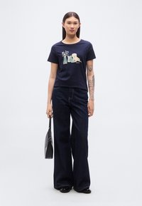 Μπλε σκούρο κοντομάνικο μπλουζάκι με γραφικό σχέδιο ενός σκύλου και λουλουδιών· συνδυασμένο με σκούρο, φαρδύ τζιν και μαύρη τσάντα.