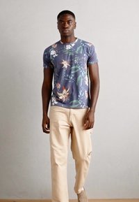 Camiseta de manga corta azul con patrón floral en tonos de rosa y verde, combinada con pantalones beige claros. Modelo de pie frente a un fondo neutral.