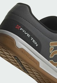 Černé tenisky s texturovaným svrškem, kontrastní bílou a gumovou podrážkou, se znakem "FIVE TEN" v červené a světlé hnědé barvě.
