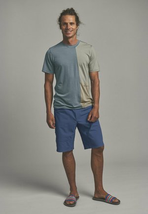 Mann mit lockigem Haar, der ein halb blaues, halb graues T-Shirt, blaue Shorts und bunte gestreifte Sandalen trägt, steht lächelnd vor einem einfarbigen Hintergrund.