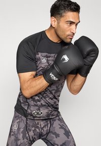 Mustast ja hallist kamuflaažist spordisärk ja retuusid, kokku sobitatud mustade poksikinnastega, millel on valged aktsendid ja brändi logo.
