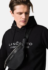 Czarna torba na pasku wykonana z fakturowanego materiału, z zamkiem błyskawicznym, regulowanym paskiem i markowymi akcentami. Noszona na czarnej bluzie z kapturem.