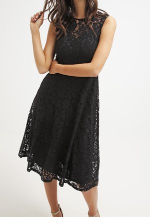 Femme portant une robe midi noire en dentelle sans manches, debout avec un bras croisé et l'autre main jouant avec ses cheveux.