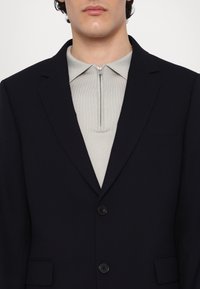 Marineblazer mit zwei Knöpfen, mit Reverskragen, kombiniert mit einem hellgrauen Zip-Neck-Top. Strukturiertes Material mit taillierter Passform und minimierter Detailgestaltung.