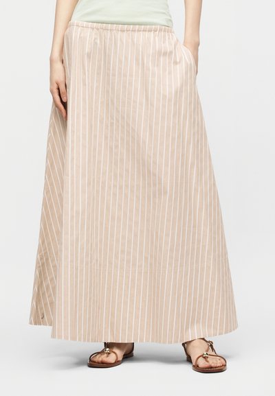 Jupe longue beige à rayures blanches verticales, portée avec des sandales plates marron et un haut vert clair rentré, mains dans les poches.